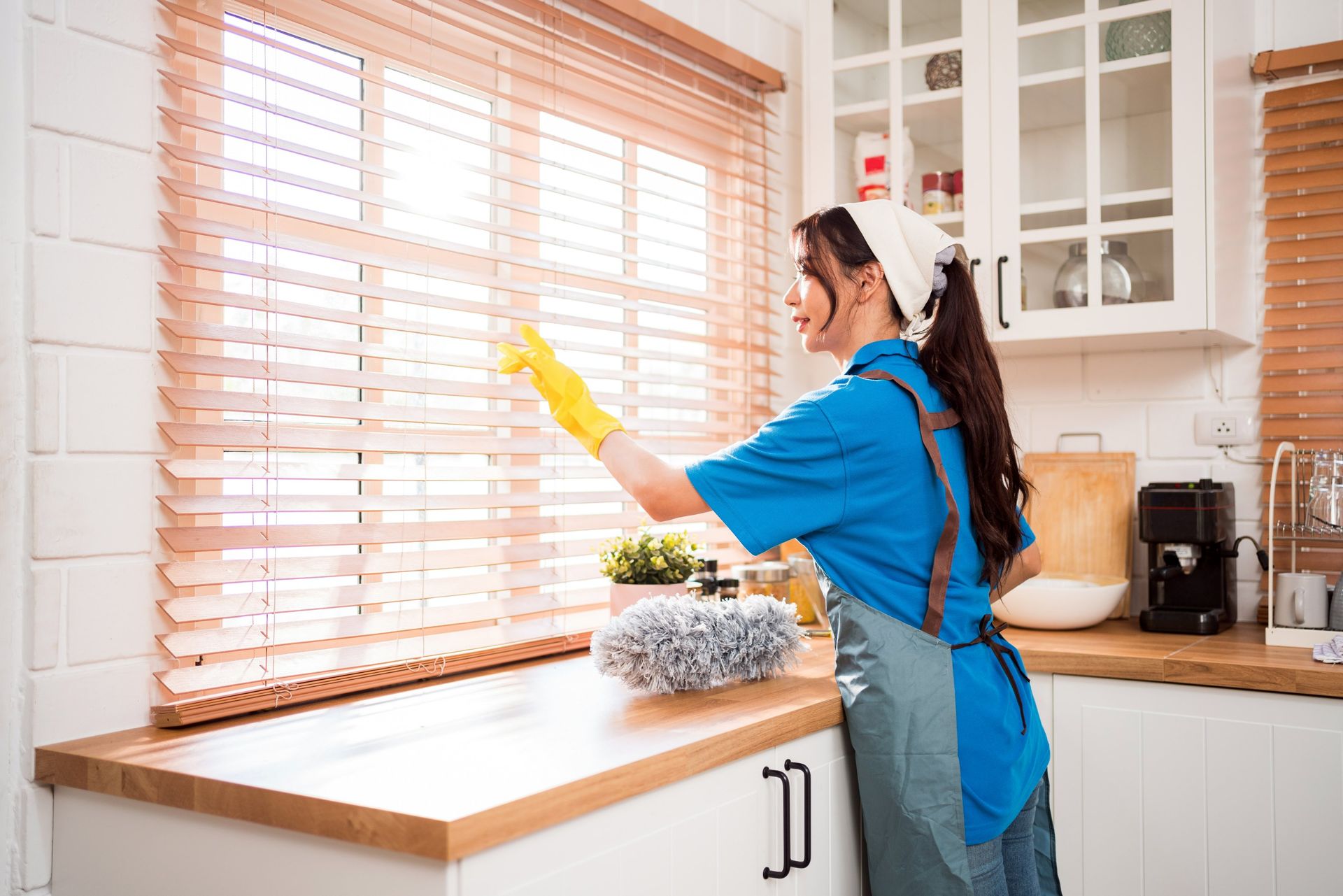 Woman in blue cleaning kitchen window, wearing gloves and apron.