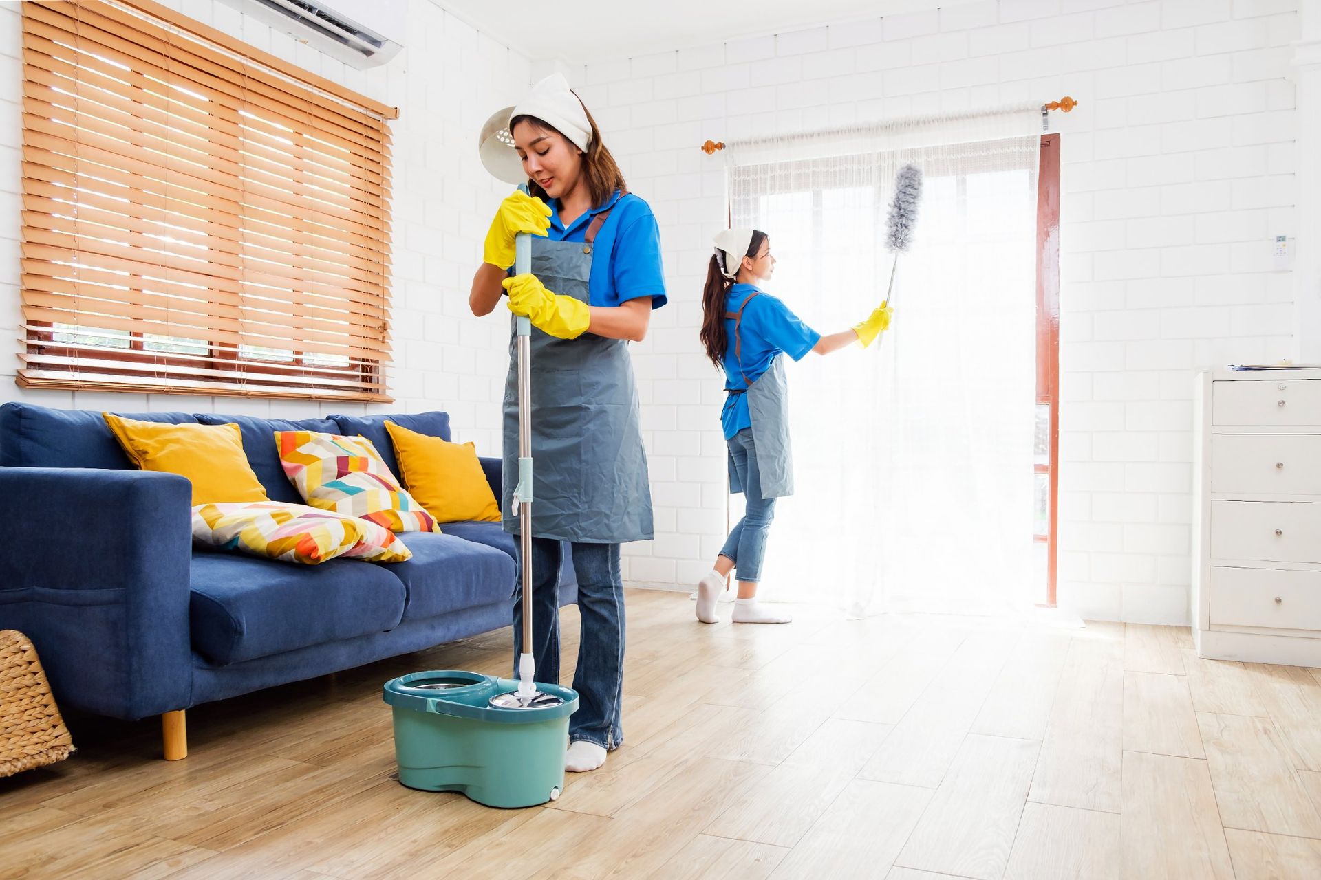 Two women cleaning a bright living room; one mopping, the other dusting a curtain.