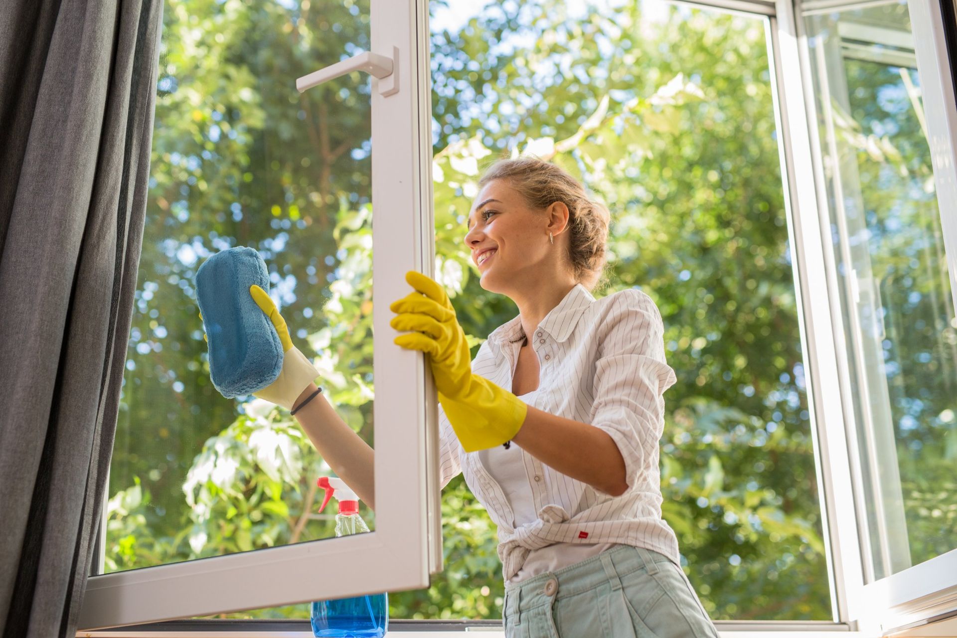 Woman cleaning window with a blue sponge, wearing yellow gloves, in front of a green outdoor backdrop.