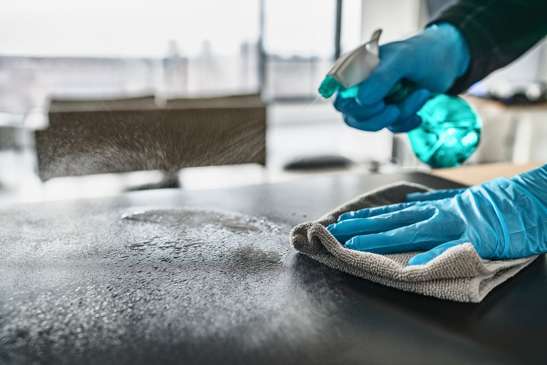 Hands in blue gloves spraying and wiping a dark surface with cleaning solution.
