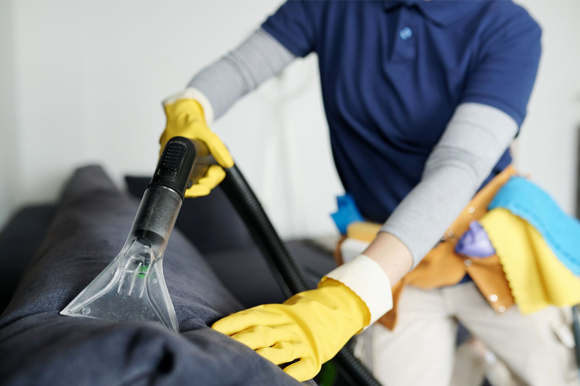 Person wearing yellow gloves vacuuming a dark gray sofa with a black handheld tool.