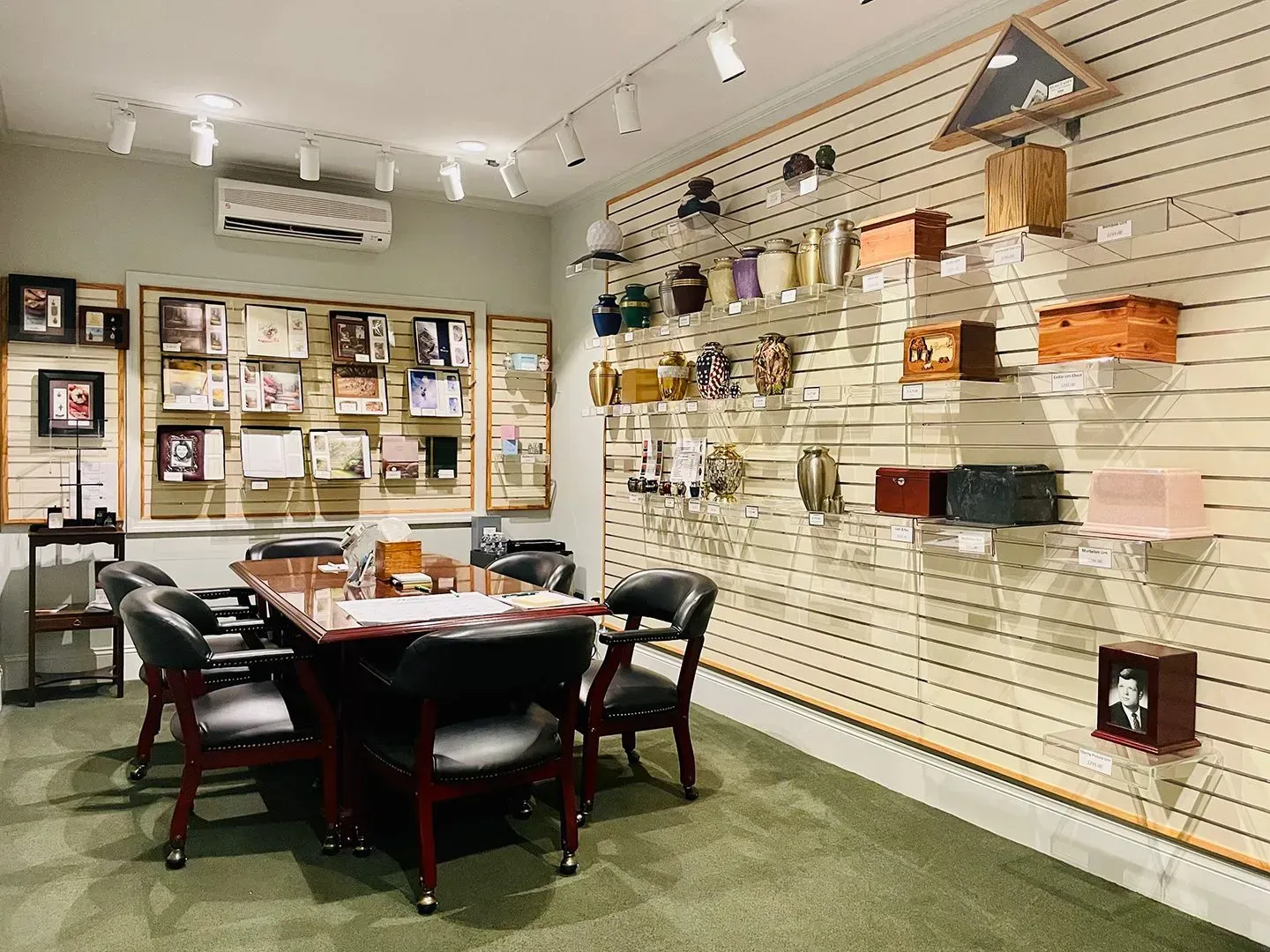 Retail showroom interior with wall displays, shelves, and a central table with chairs on green carpet.