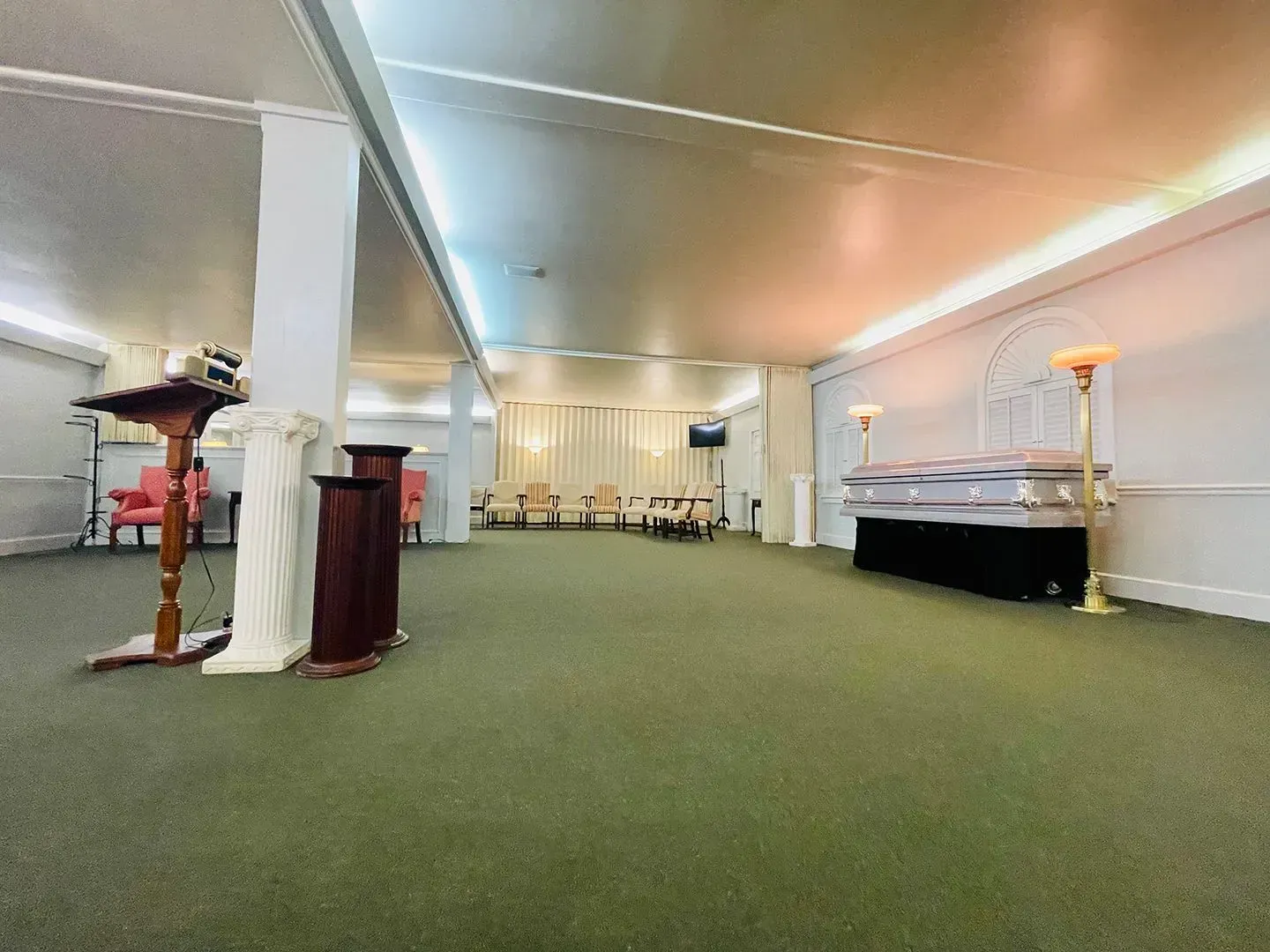 Spacious indoor room with green carpet, white walls, and scattered furniture under warm lights.