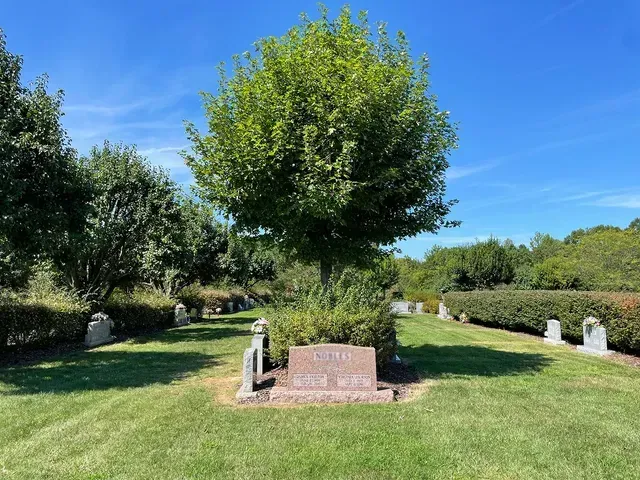 Gravestone in a sunny cemetery with green grass, trees, and a bright blue sky