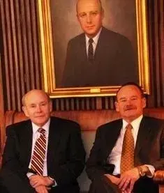Two men seated in suits beneath a large framed portrait in a wood-paneled room