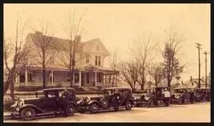 Sepia photo of early 1900s cars parked along a street in front of a large house