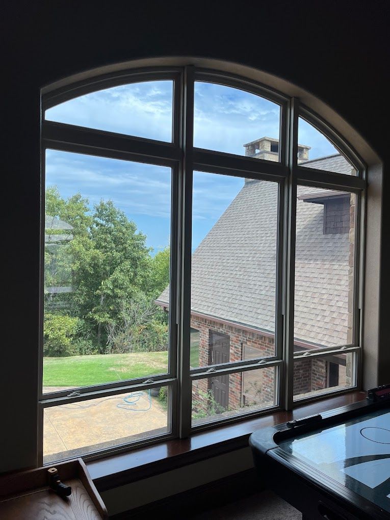 Large arched window overlooking a house with a brick exterior, green trees, and a cloudy blue sky.