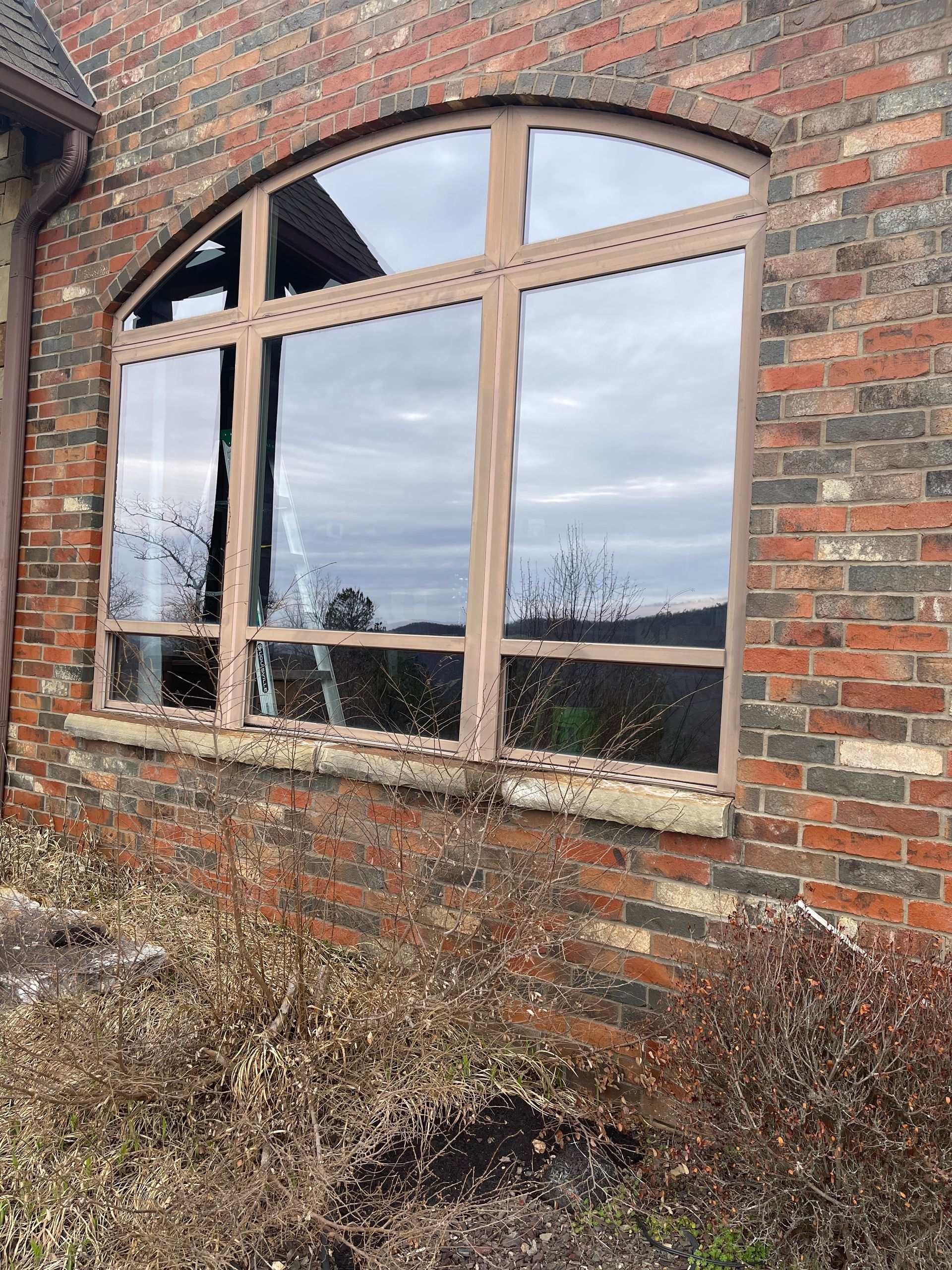 Large arched window in a brick building reflects a cloudy sky and dry bushes.
