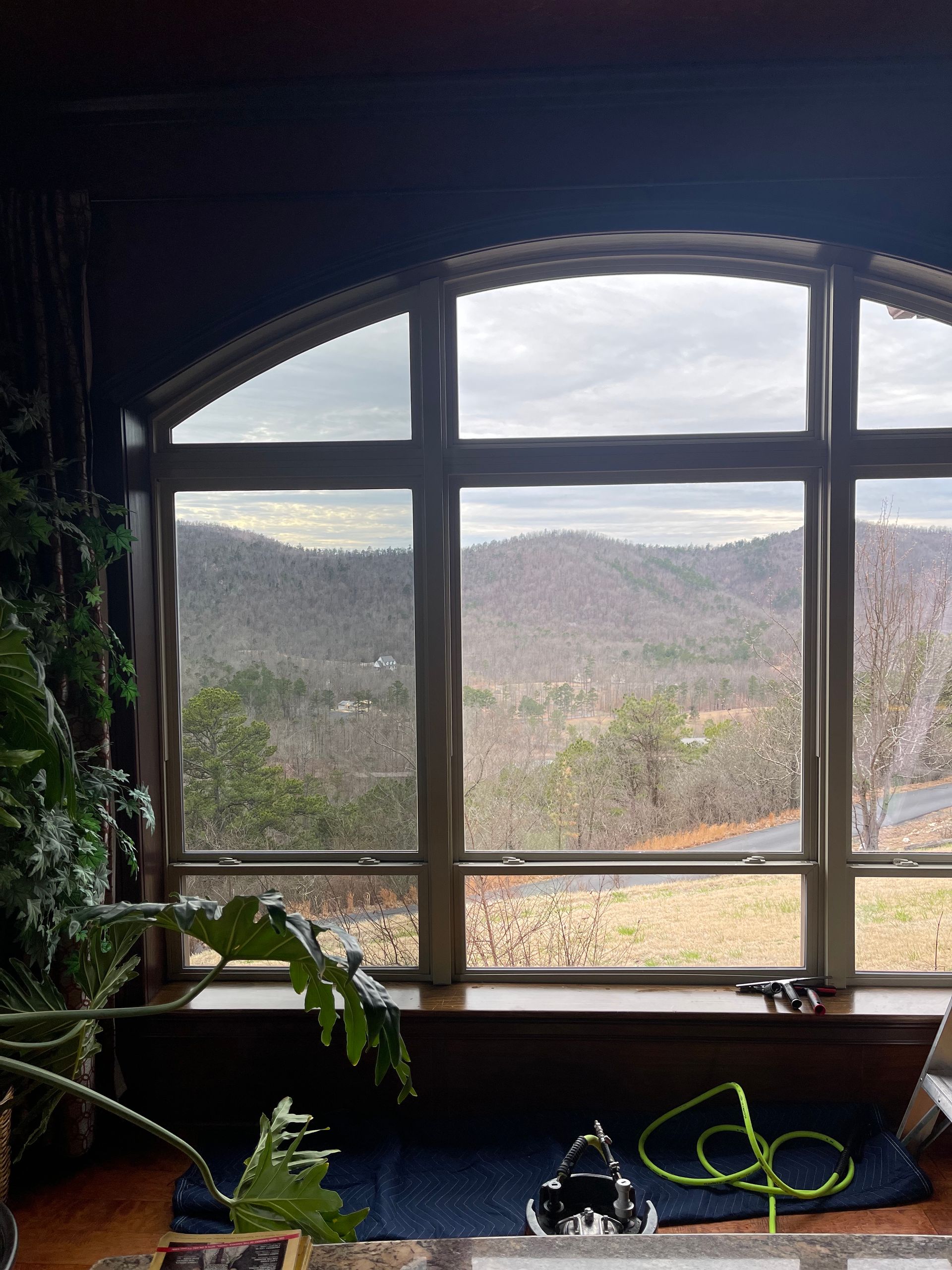 Arched window framing a mountain view on a cloudy day. Inside, a built-in bench with plants.