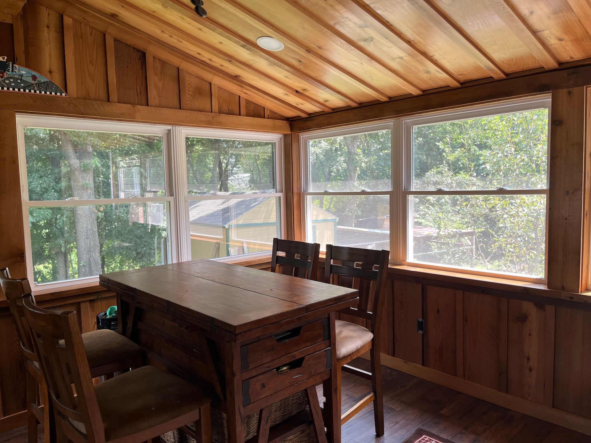 Wood-paneled sunroom with windows, table, and chairs. Sunlight streams through the windows, illuminating the interior.