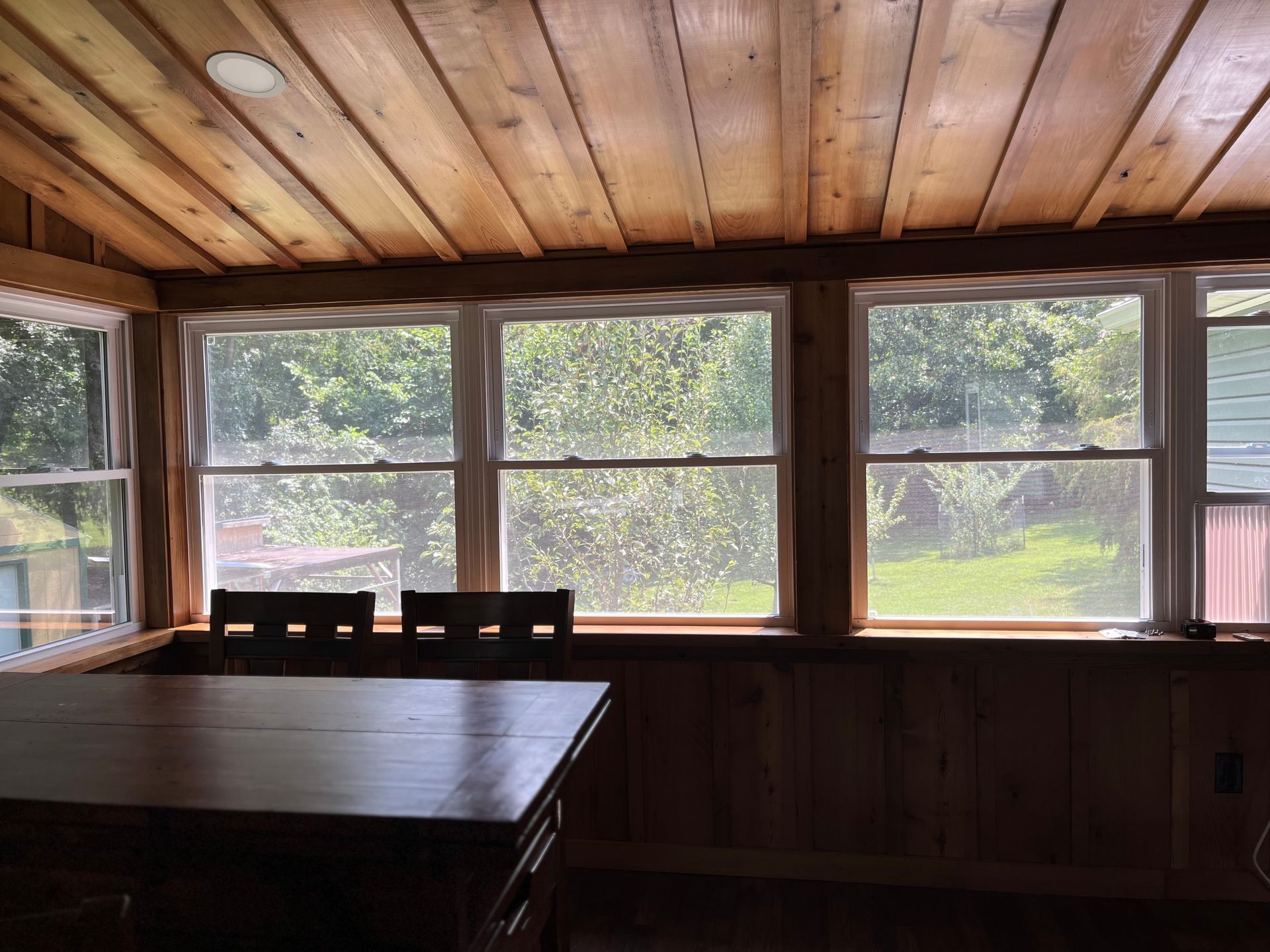 Wooden-paneled sunroom with large windows overlooking trees; a dark wood table with three chairs is inside.