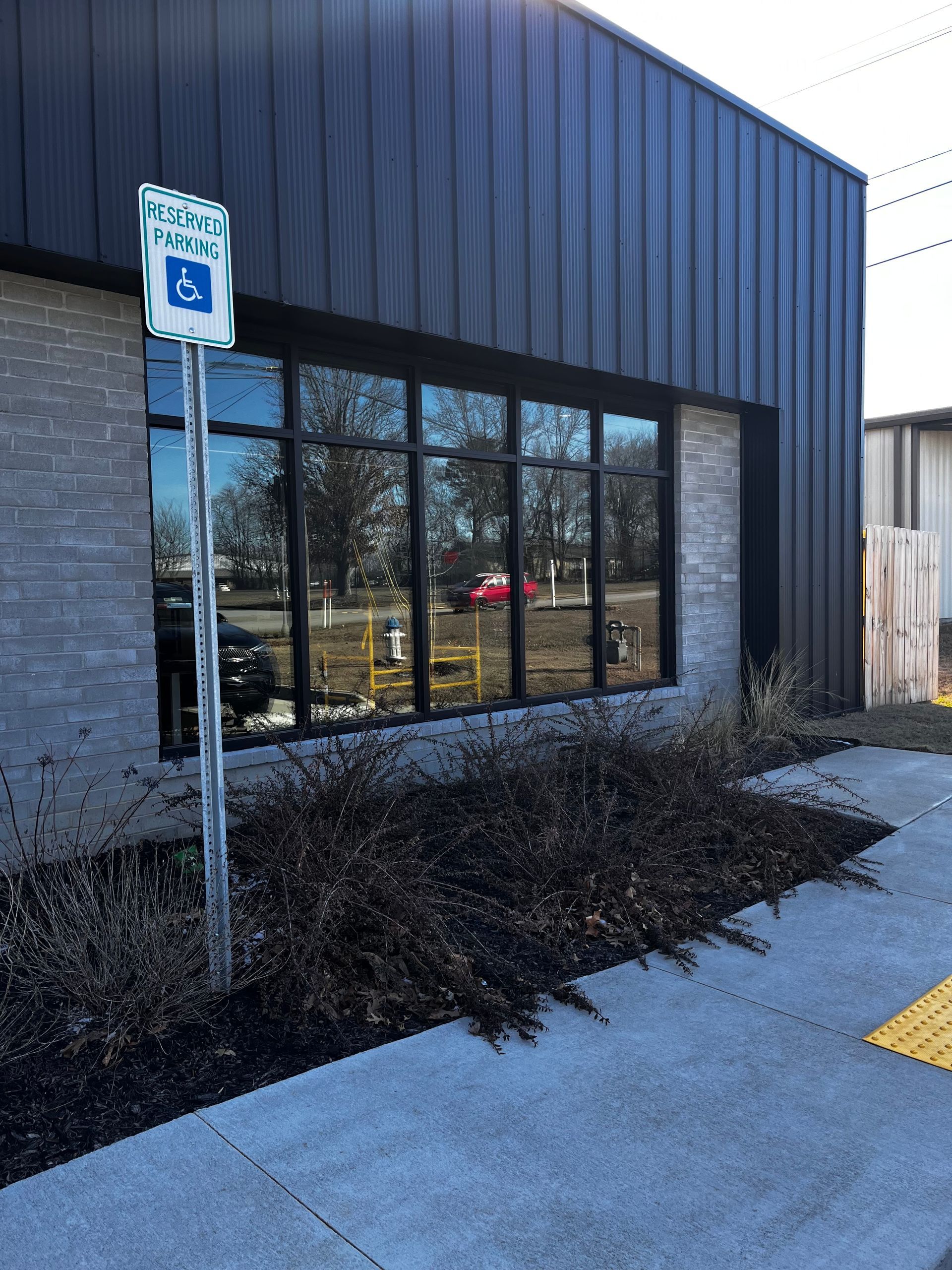 Building exterior with accessible parking sign. Windows reflect trees, with dry bushes and sidewalk in the foreground.
