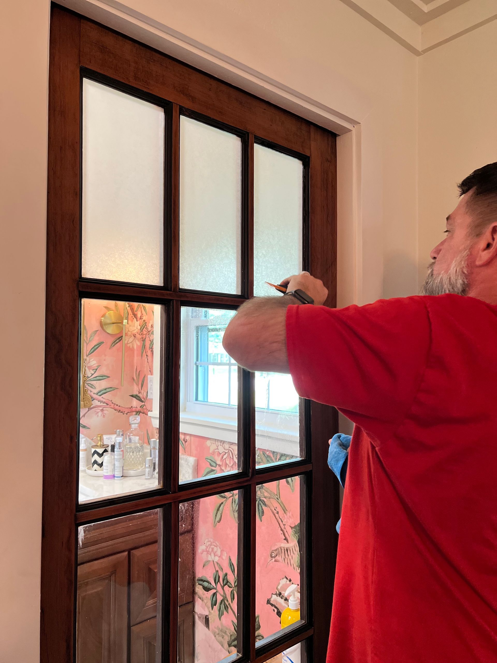 Man in red shirt adjusts window panes with frosted glass in a room with floral wallpaper.