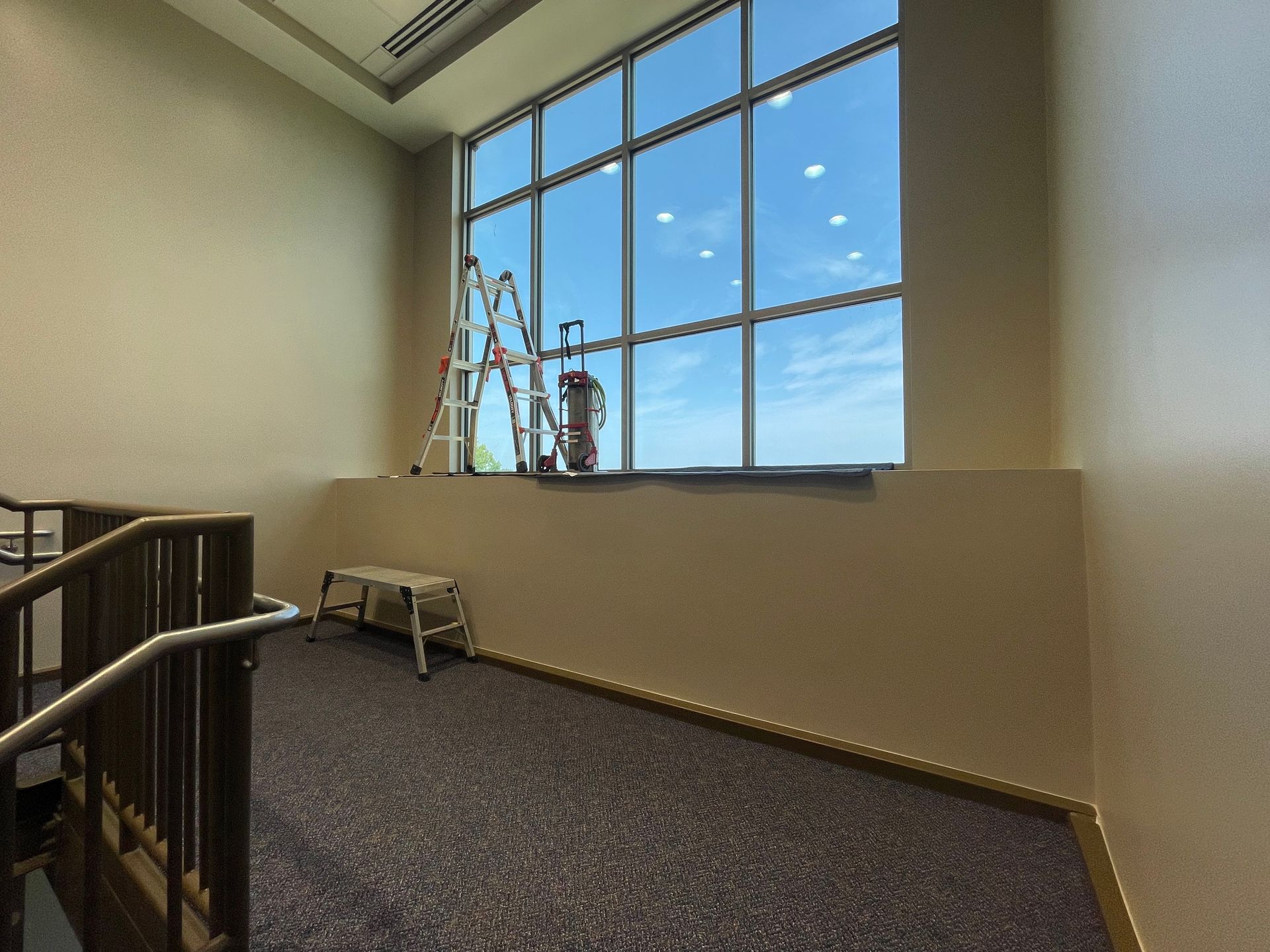 A long window with a ladder and step stool. Interior hallway with a dark carpet and blue sky visible.