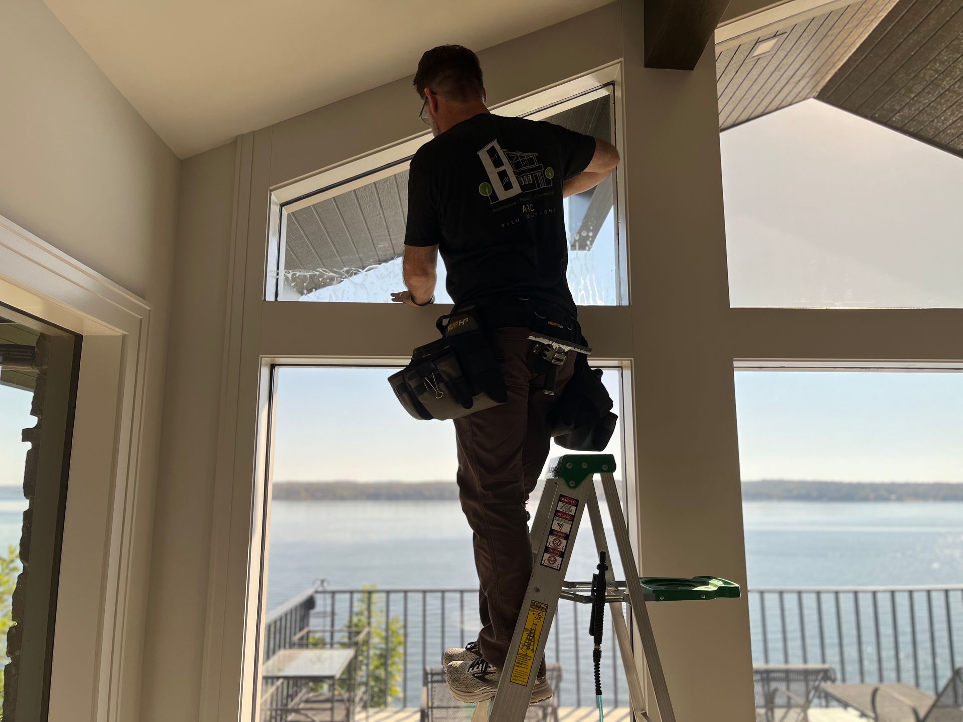 Person on a ladder applying tint to a large window overlooking a lake.