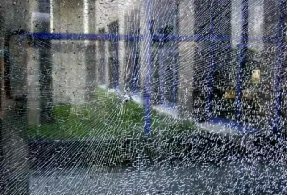 Shattered glass window with a web of cracks, reflecting a blurred outdoor scene with greenery and buildings.