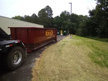 Red dumpster beside a driveway and grassy verge near trees and utility bins