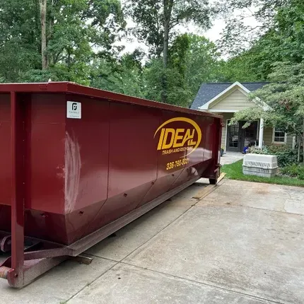 Maroon dumpster in a driveway beside a house, with green trees in the background.