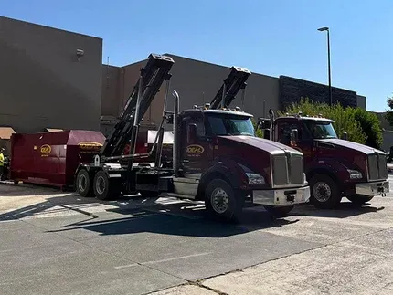 Maroon semi-truck with boom lift and red dumpster parked in a sunny lot