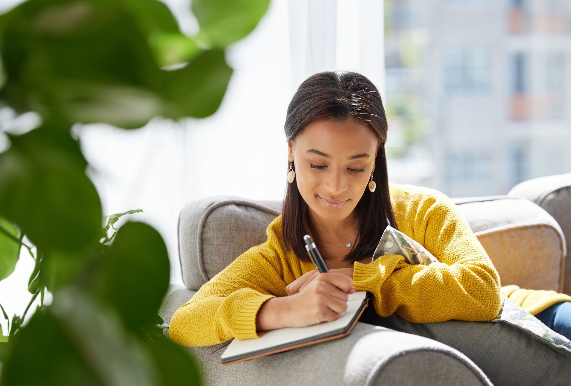 Woman in yellow sweater writing in a notebook, sitting on a gray couch in a room with a plant.