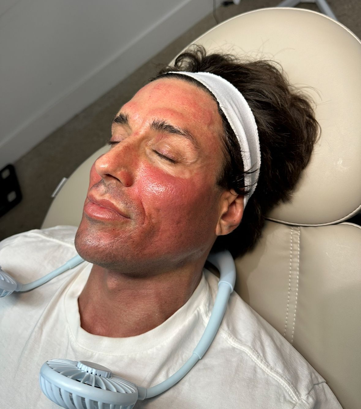 Person reclining in a chair with a red facial mask and a white headband in a treatment room