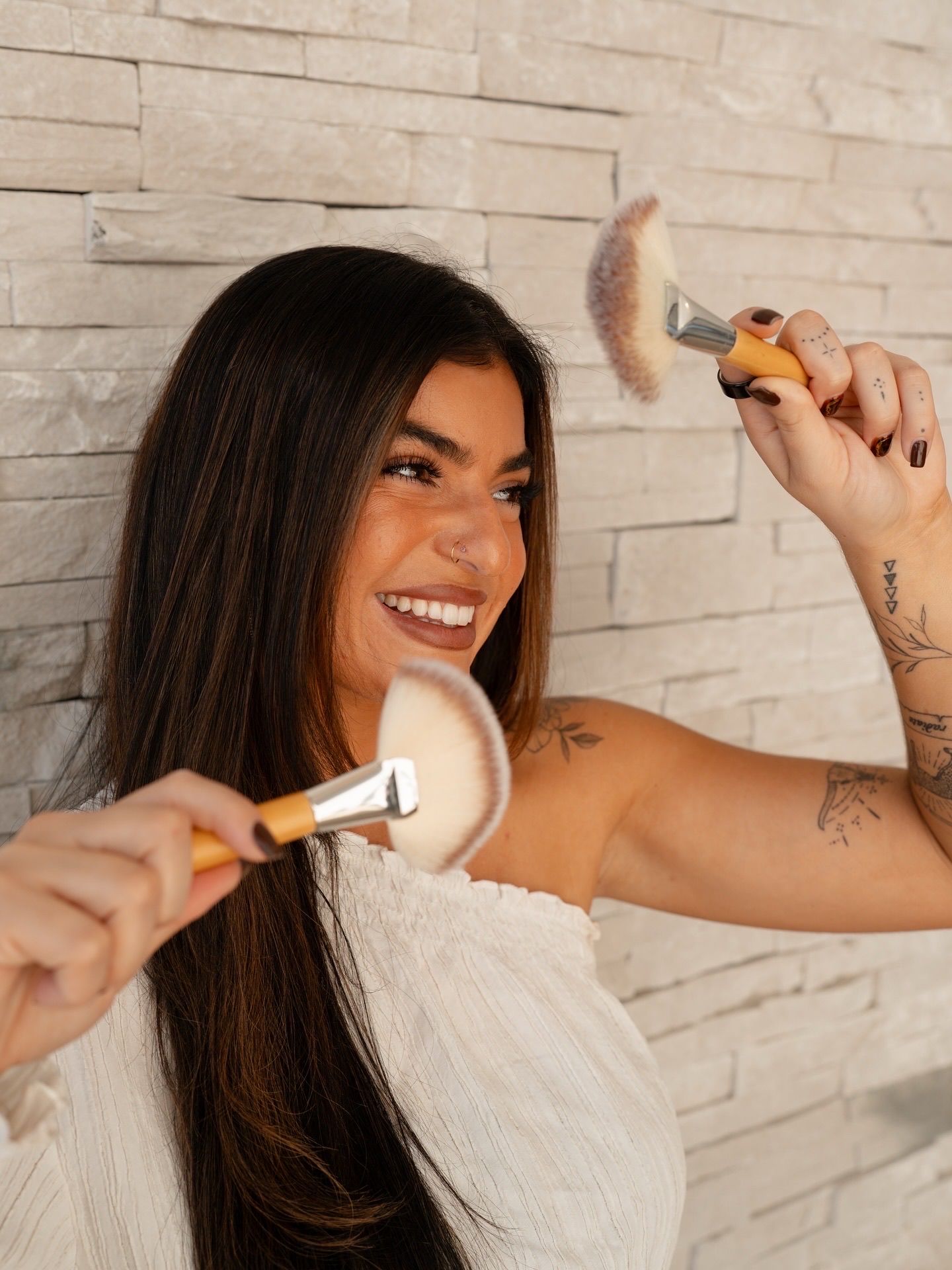 Woman smiling, holding makeup brushes, white top, against a textured wall.