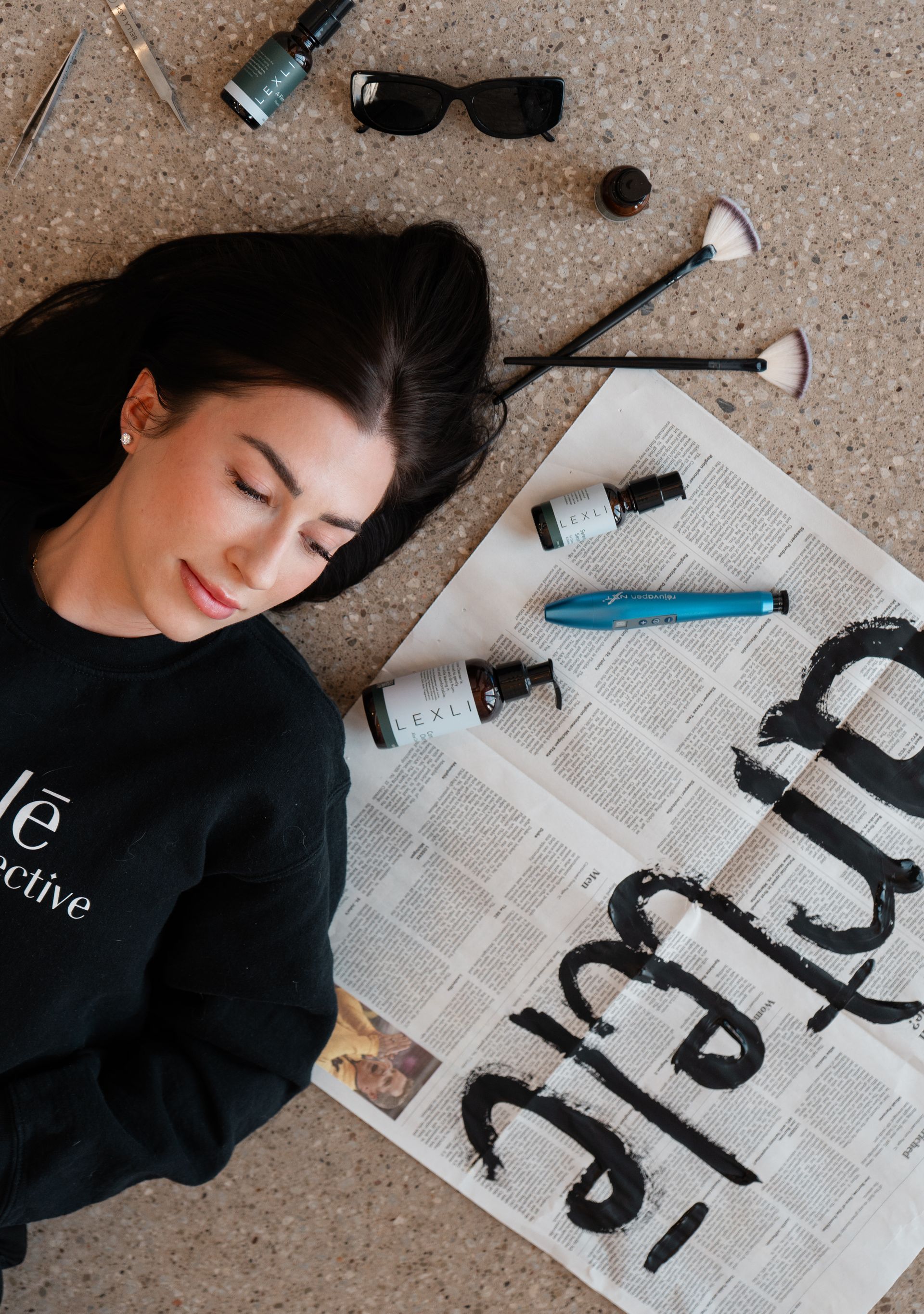 Woman lying on carpet beside newspapers, brushes, sunglasses, and paint supplies, wearing a black shirt.