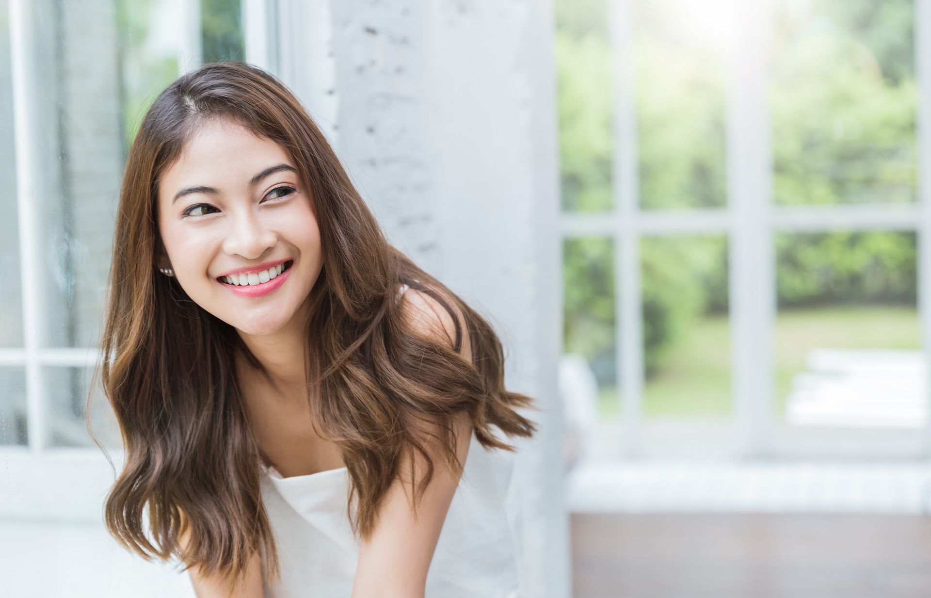 A woman in a white dress is smiling in front of a window.