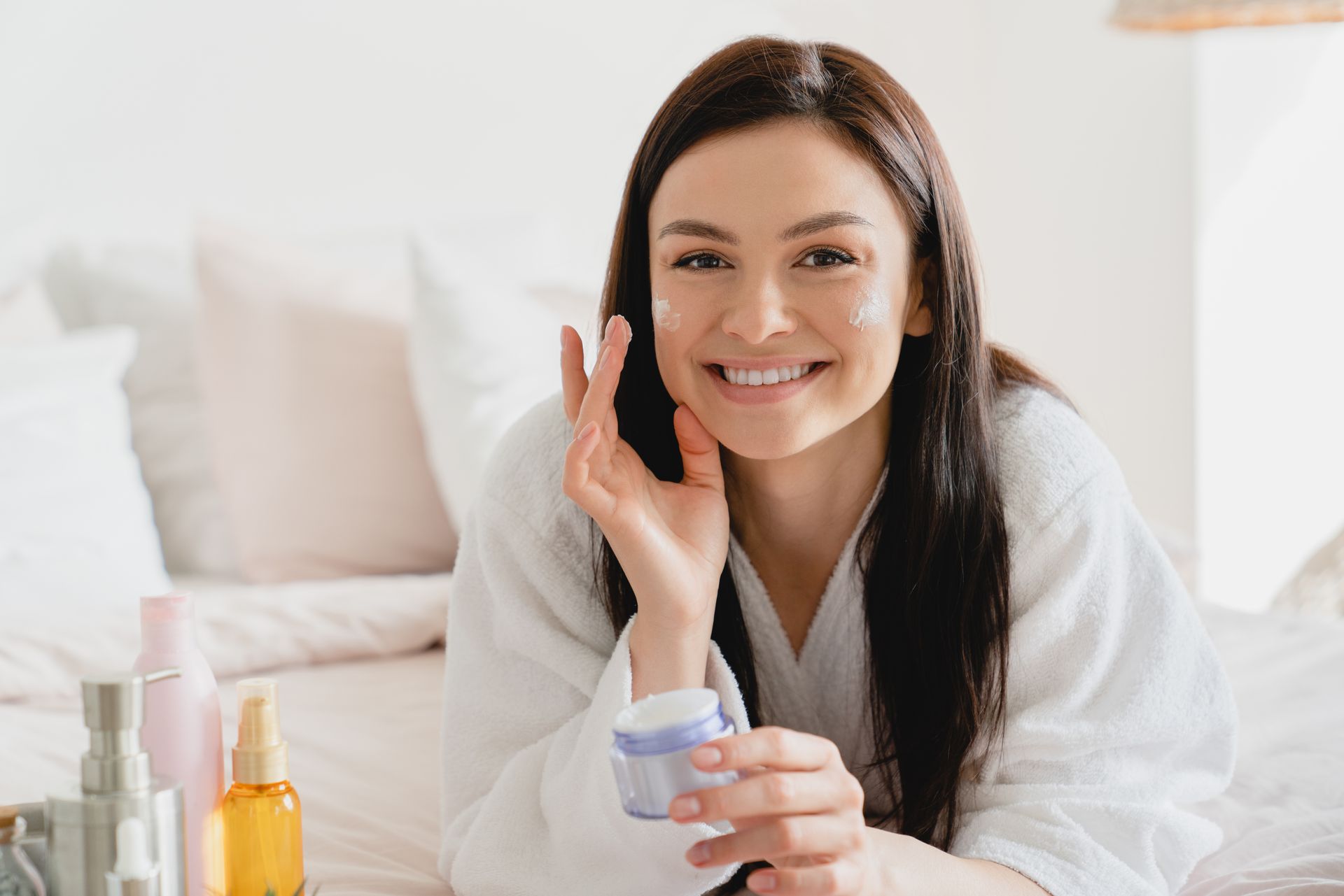 A woman is laying on a bed applying cream to her face.
