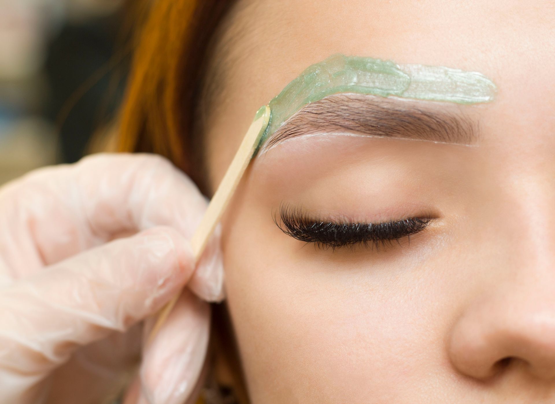 Gloved hand applying green eyebrow wax to a closed eye with makeup