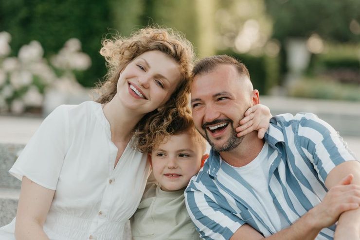 A smiling couple and their young child pose closely together outdoors in a sunlit setting.
