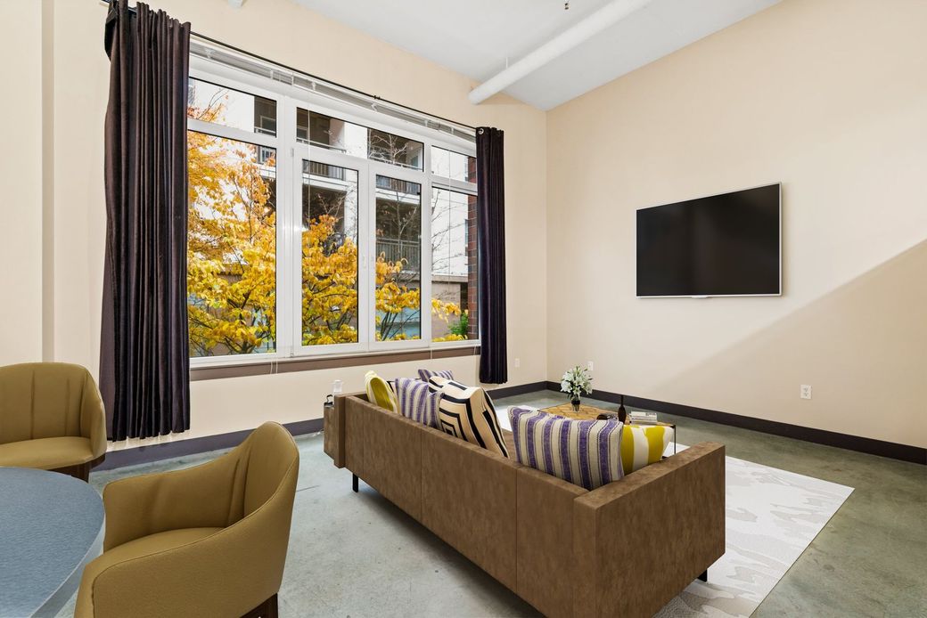 Modern living room with green sofa, fireplace, large wall TV, and wooden chairs near bright windows.