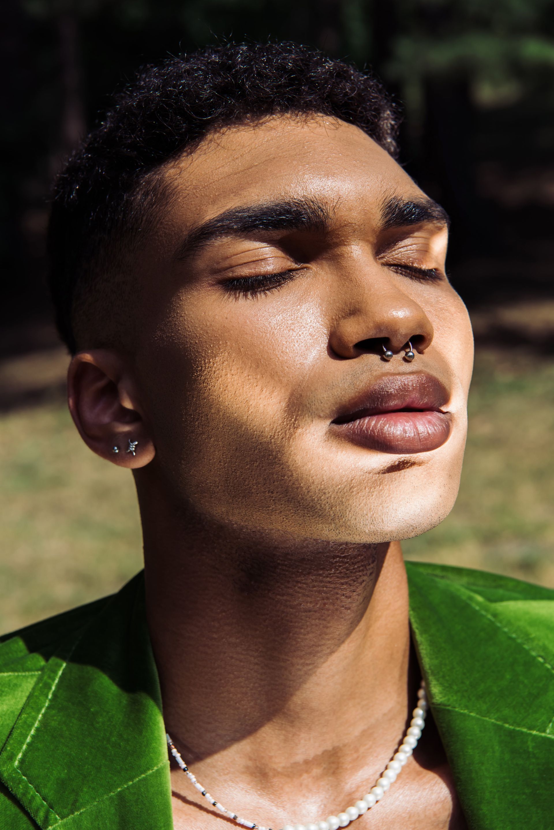 A close up of a person wearing a green jacket and a pearl necklace with their eyes closed.