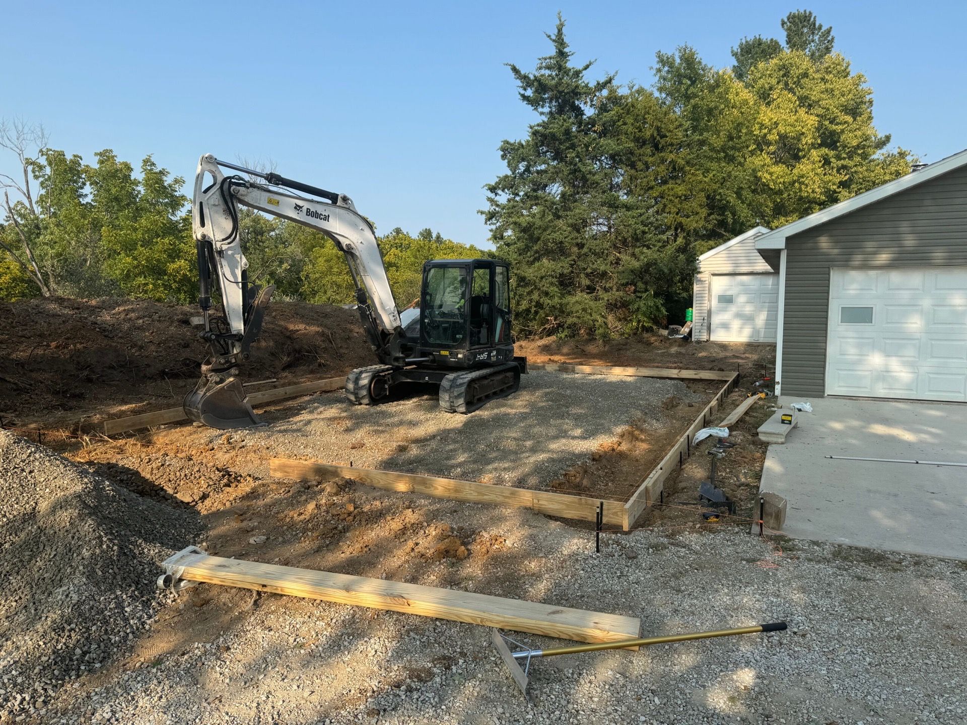 Excavator leveling dirt in a residential driveway beside a garage and trees