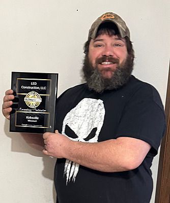 Smiling person in a black shirt and cap holding a framed award plaque against a plain wall
