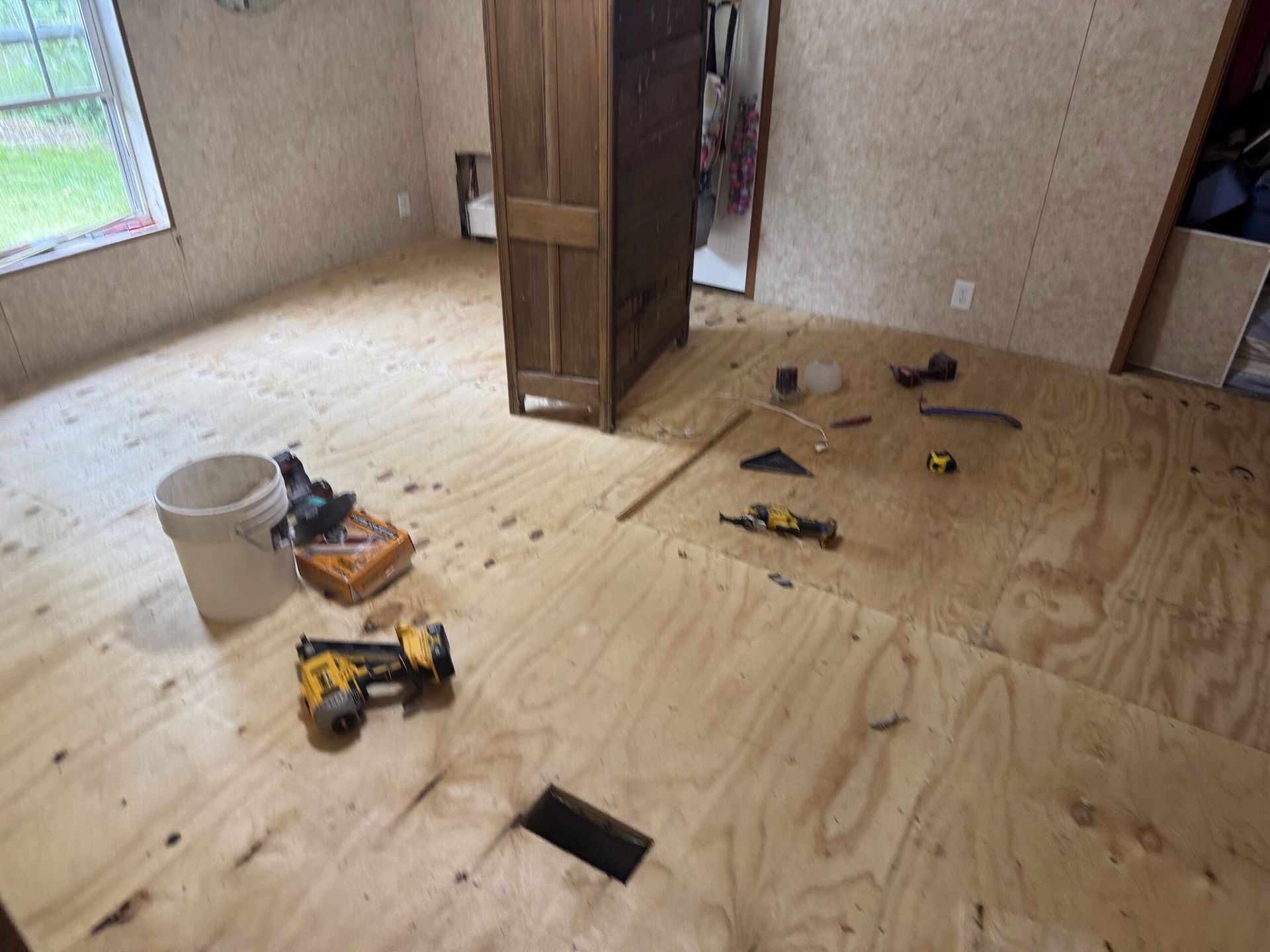 Plywood-floored room under renovation with tools, a bucket, and scattered materials.