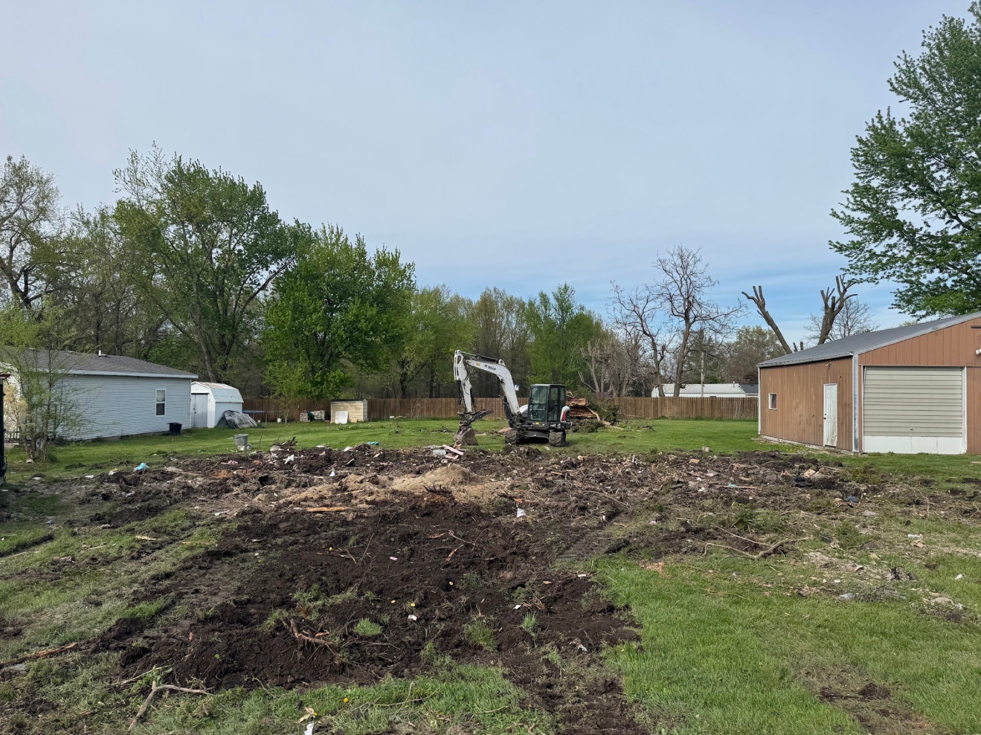 Small excavator clearing muddy yard beside a garage and mobile home under cloudy skies