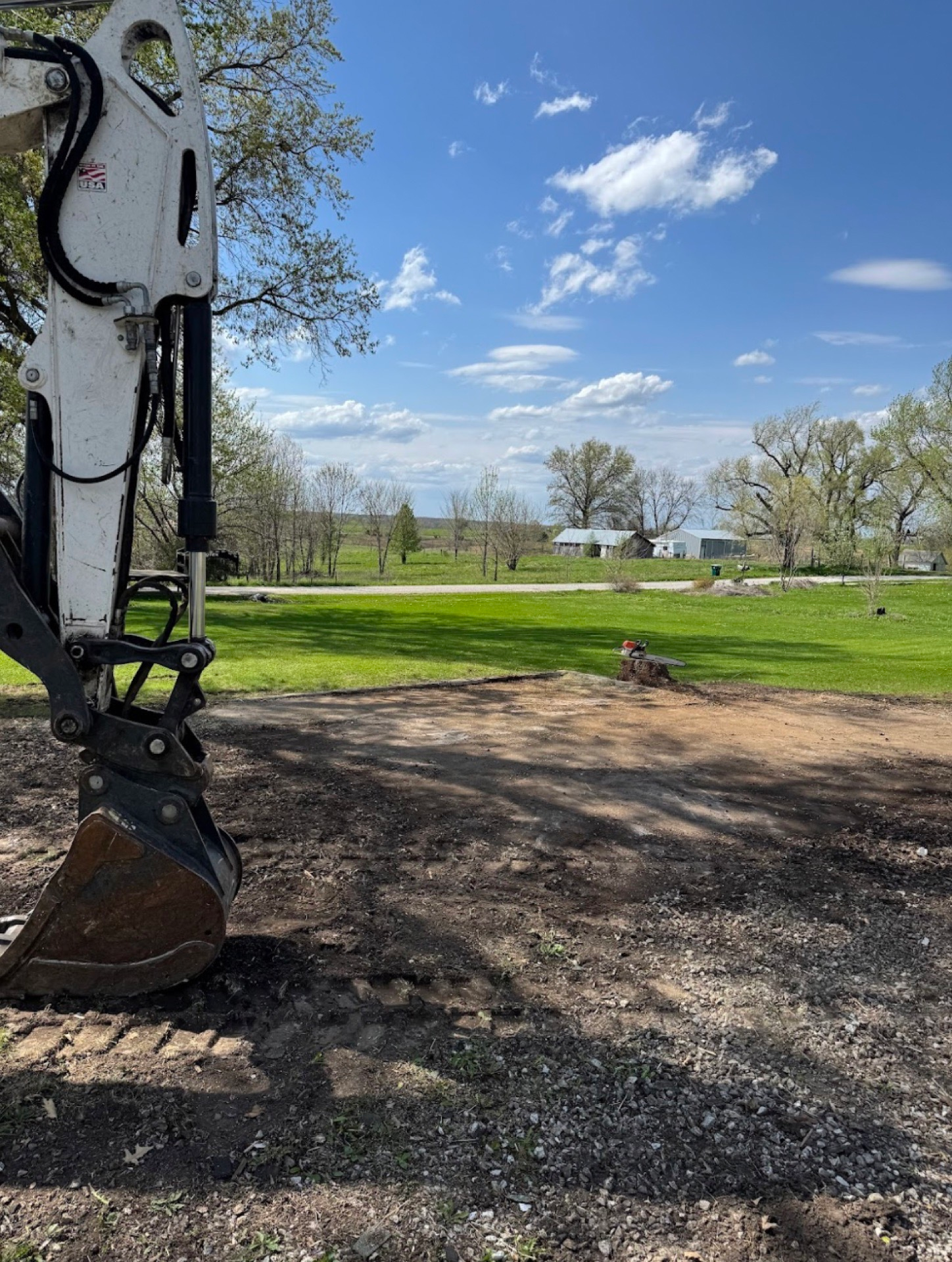 Excavator bucket at left beside a dirt area, with green field, trees, and blue sky with clouds in the background