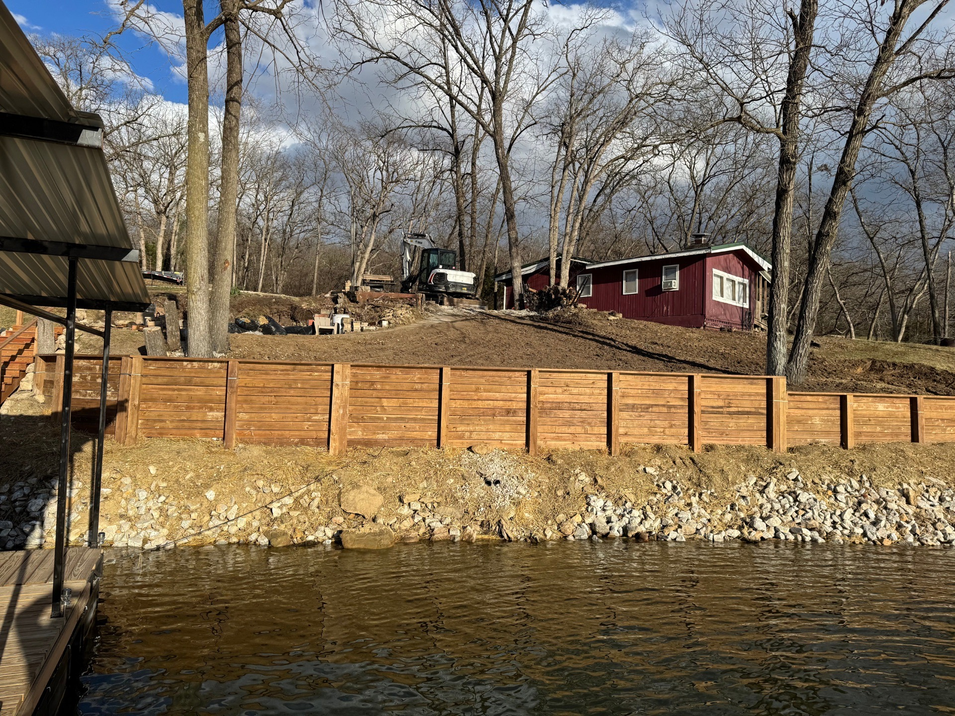 Wooded lakeside with a stone seawall, wooden fence, and red cabin under a partly cloudy sky