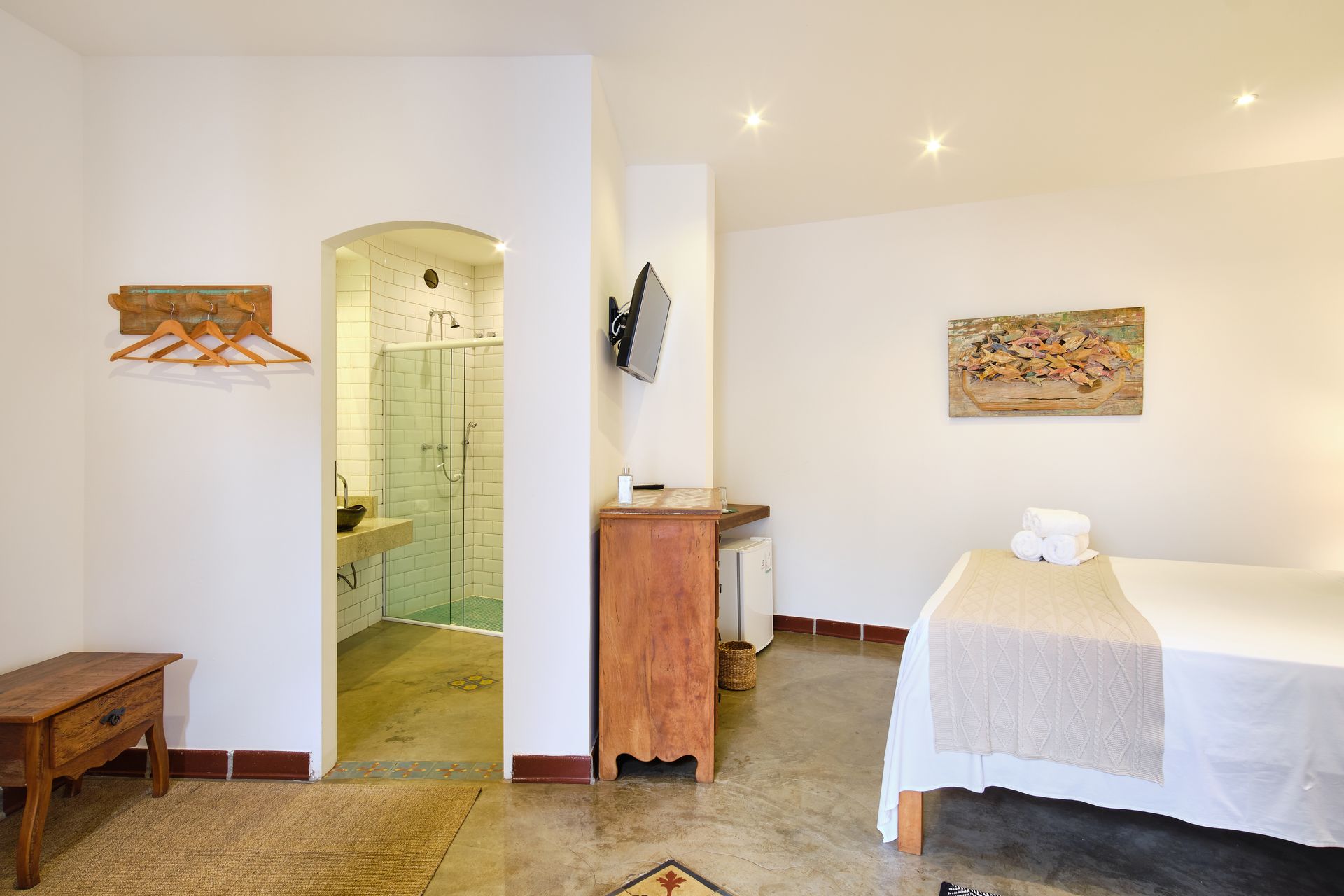 A bedroom with a bed, wood dresser, wall-mounted TV, and an arched doorway leading to a tiled bathroom with a shower.