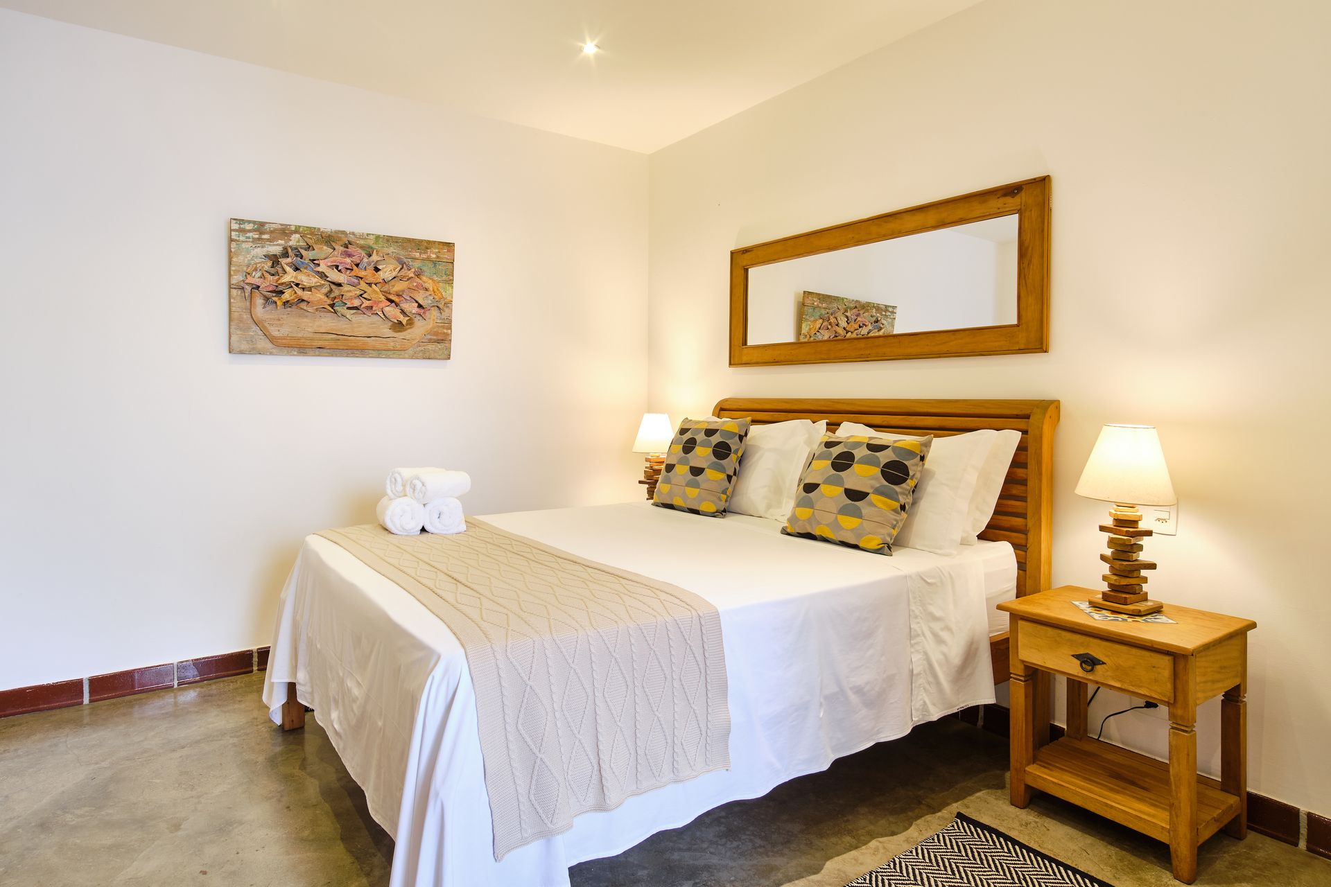A tidy bedroom featuring a queen bed with white linens, a patterned runner, two accent pillows, and a wooden nightstand.