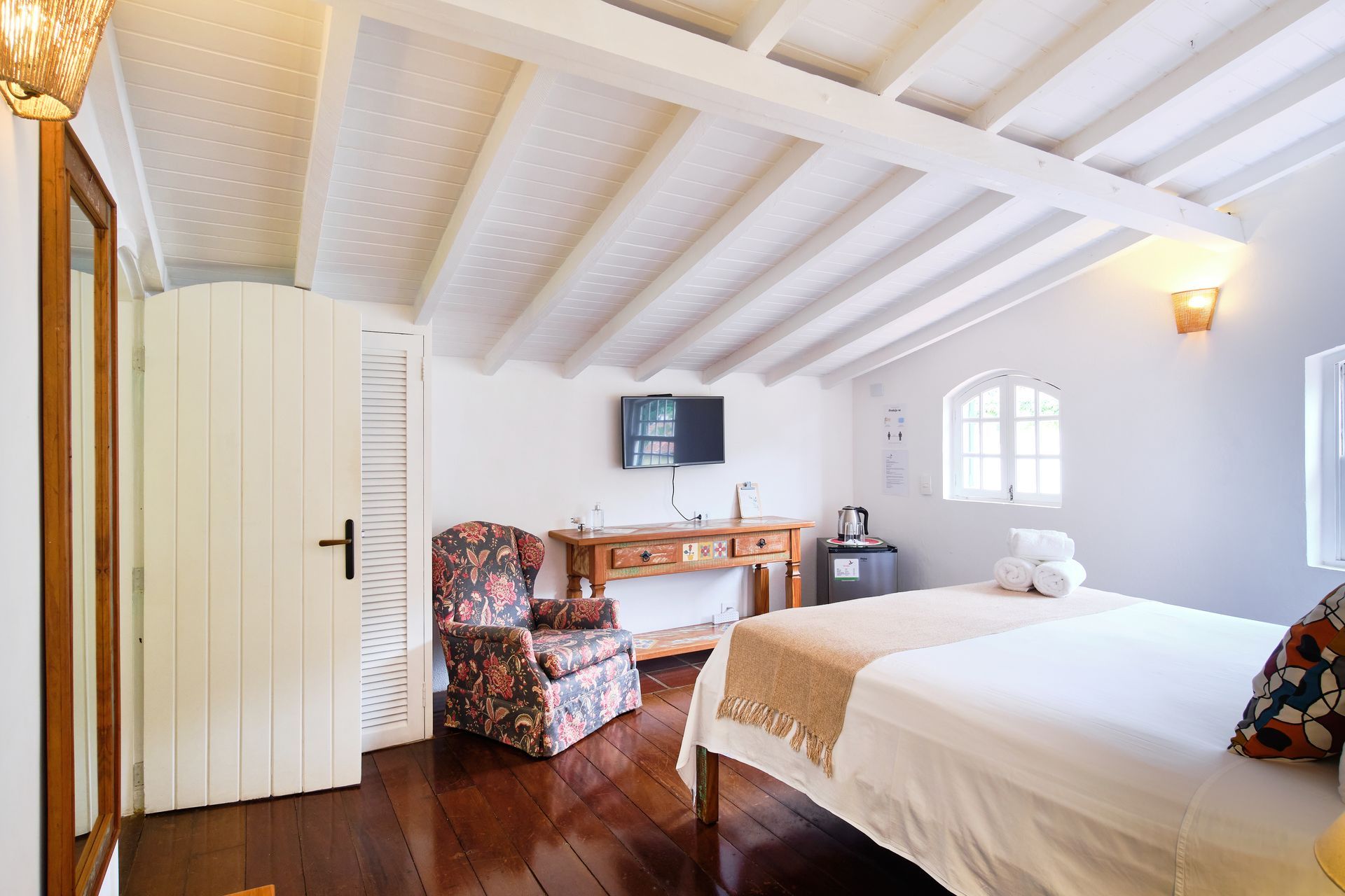 Bright attic bedroom with white beams, bed, desk, floral chair, and small arched window.