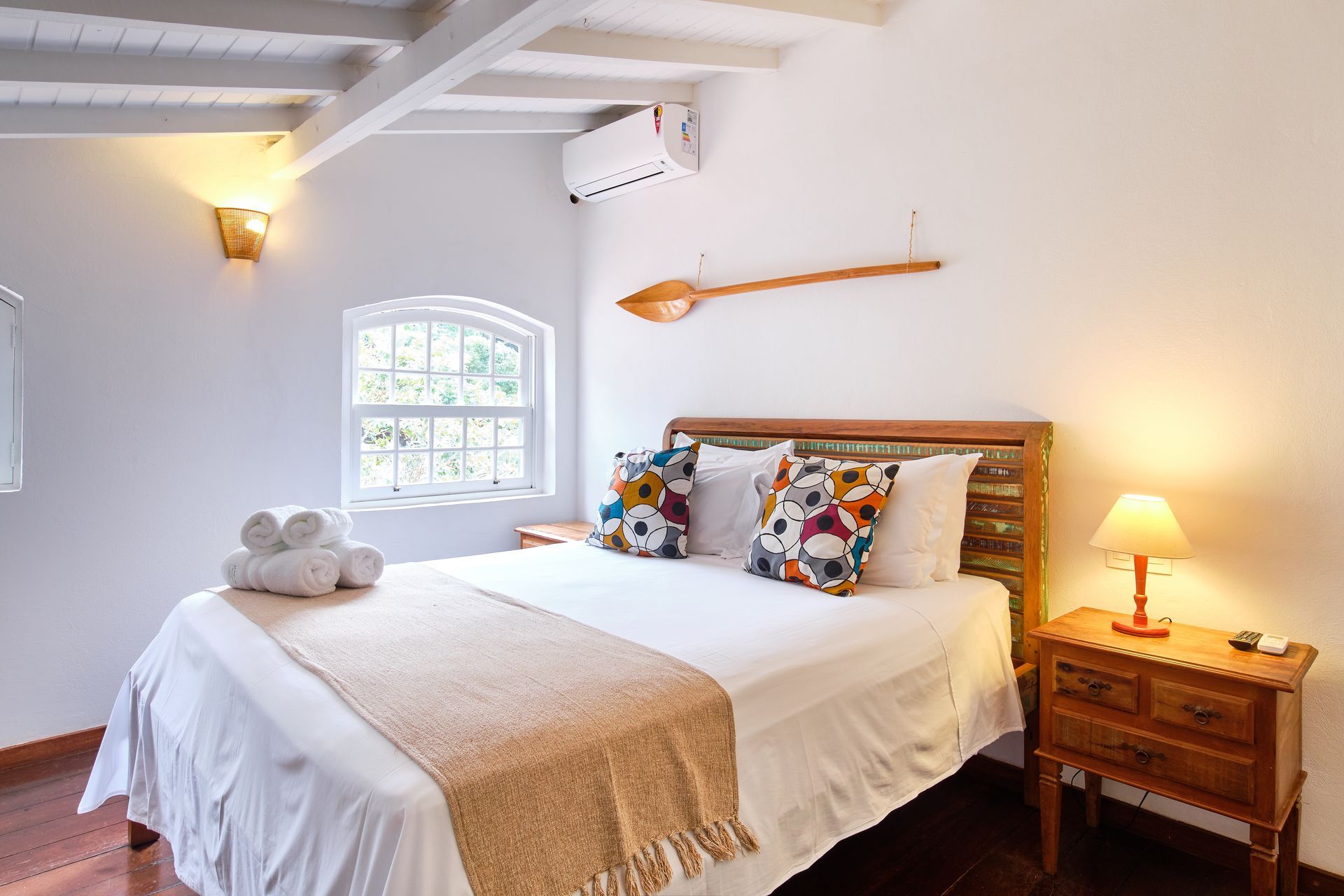 A cozy bedroom with a white-walled interior, a bed with a wooden headboard, decorative pillows, and a wall-mounted oar.