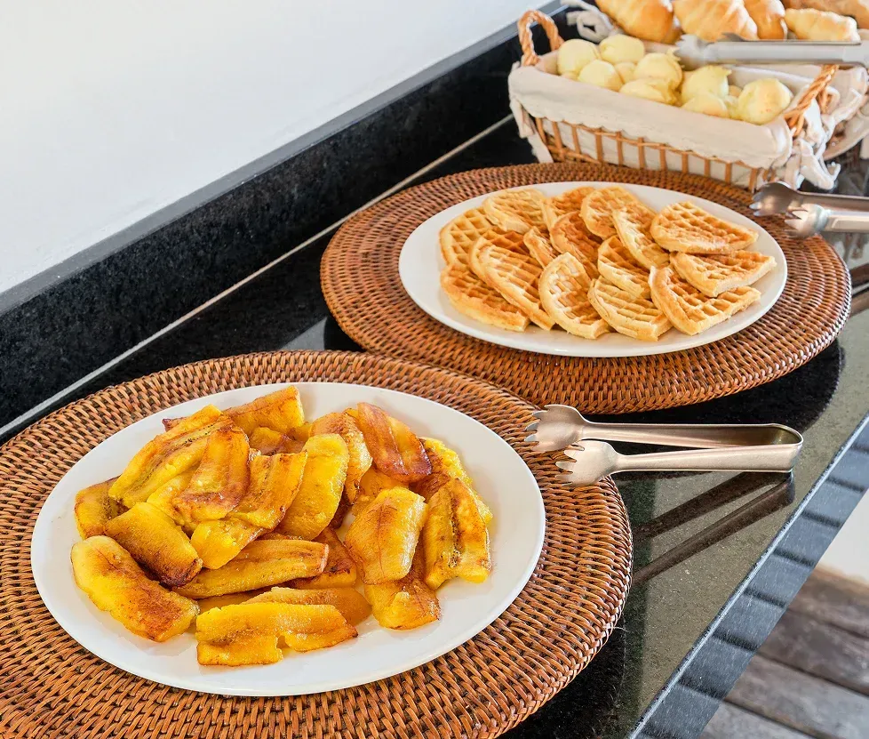Plates of fried plantains and pastries on woven placemats in a kitchen countertop setting
