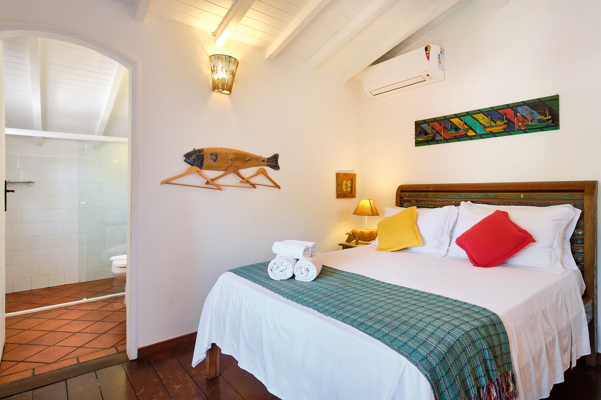 A hotel room with a queen bed featuring a green runner, a red pillow, a yellow pillow, and a bathroom visible through a door.