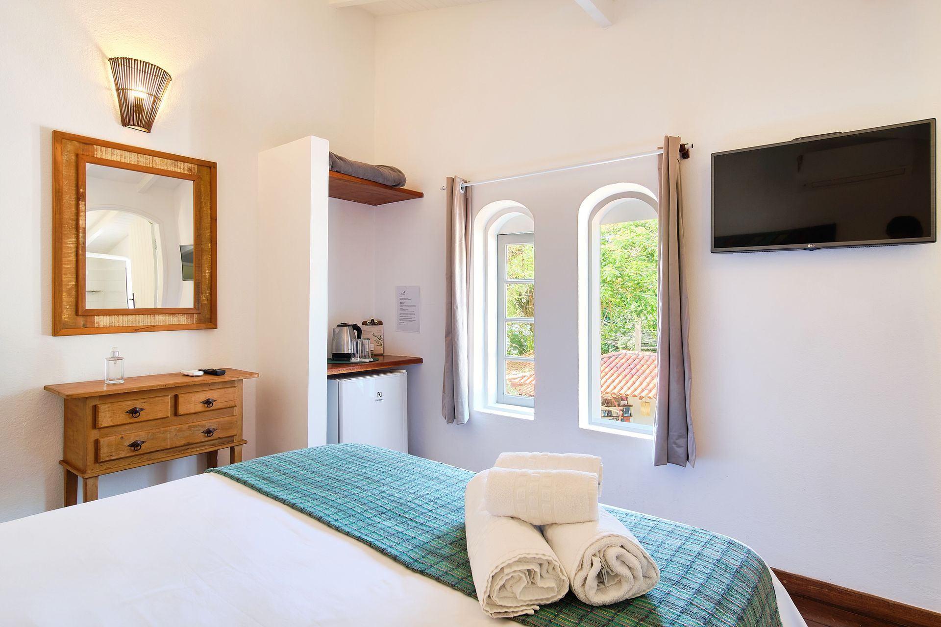 A hotel room with a bed featuring a teal runner and towels, a wooden dresser with a mirror, and a wall-mounted television.