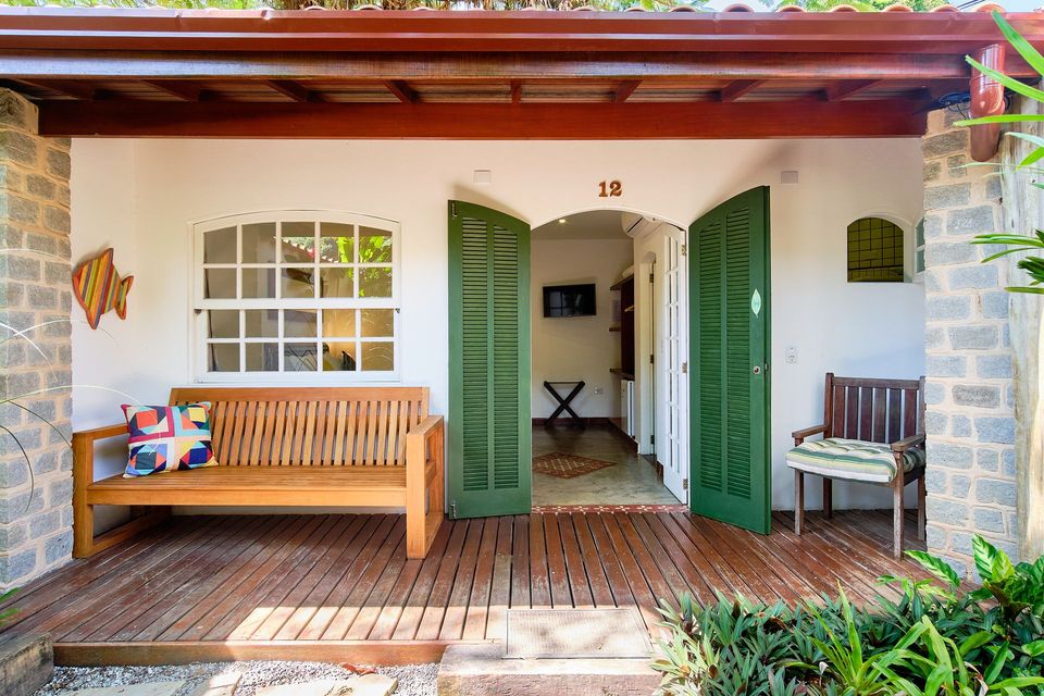 A wooden bench and a chair on a covered porch with green doors leading into a bright room.
