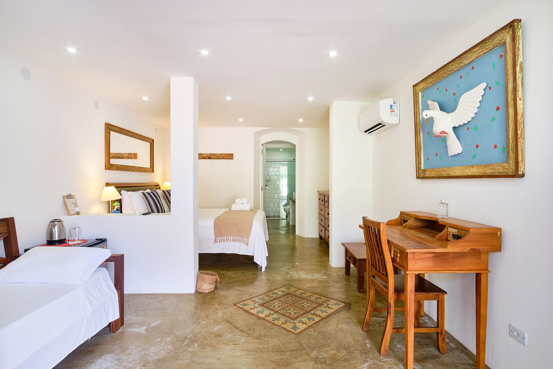 A spacious, bright hotel suite with a bed, a wooden desk with a chair, and a decorative framed dove artwork on the wall.