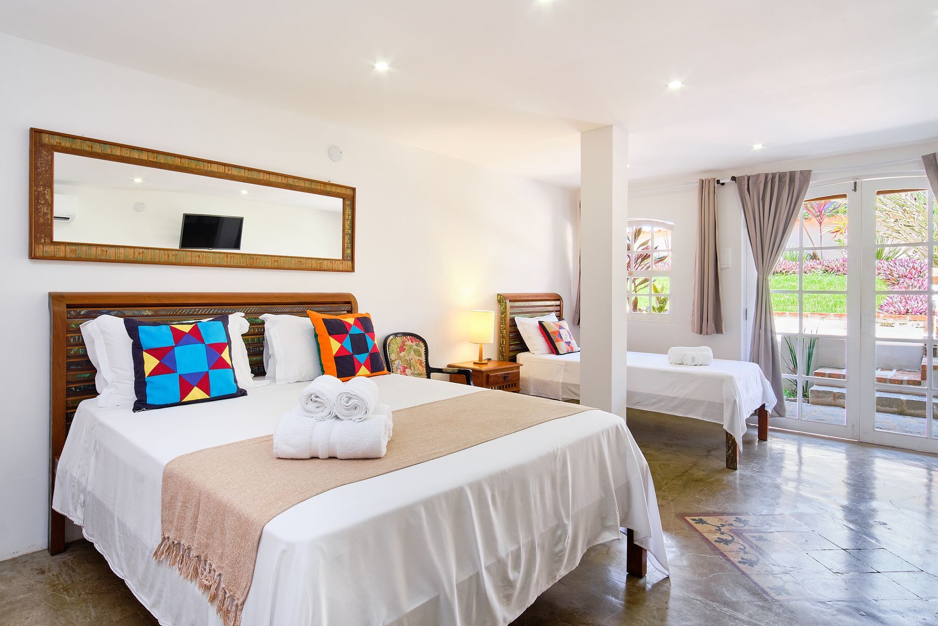 Bright bedroom with king bed, colorful pillows, mirror, and balcony doors in a modern suite