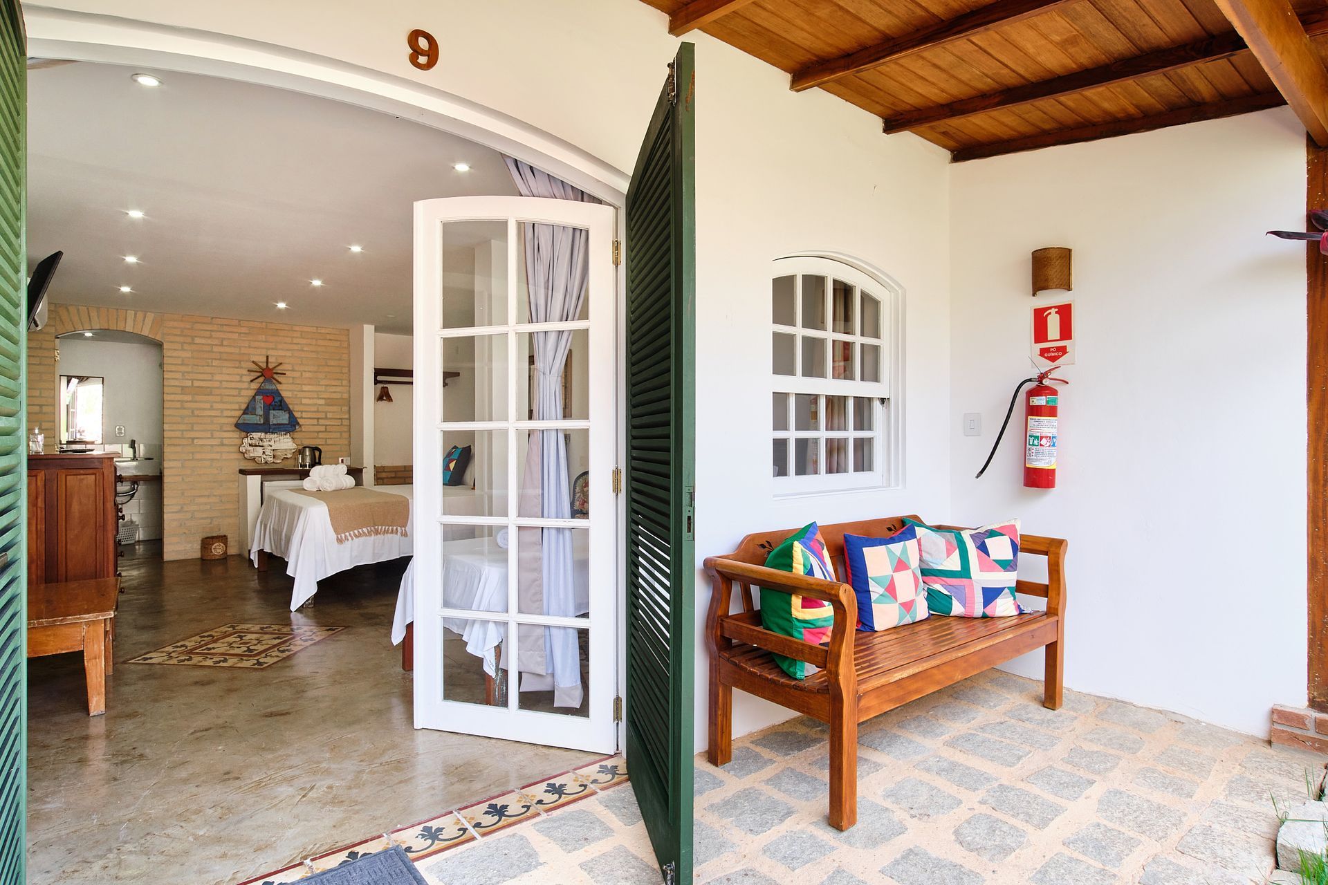 A wooden bench with colorful cushions sits on a tiled patio next to an open doorway leading into a guest room.